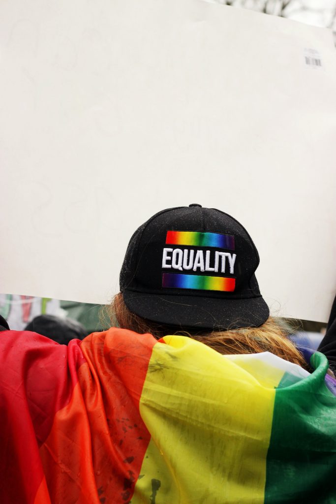 A photograph of someone from behind with a pride flag on their shoulders, wearing a cap backwards with the word "Equality" on