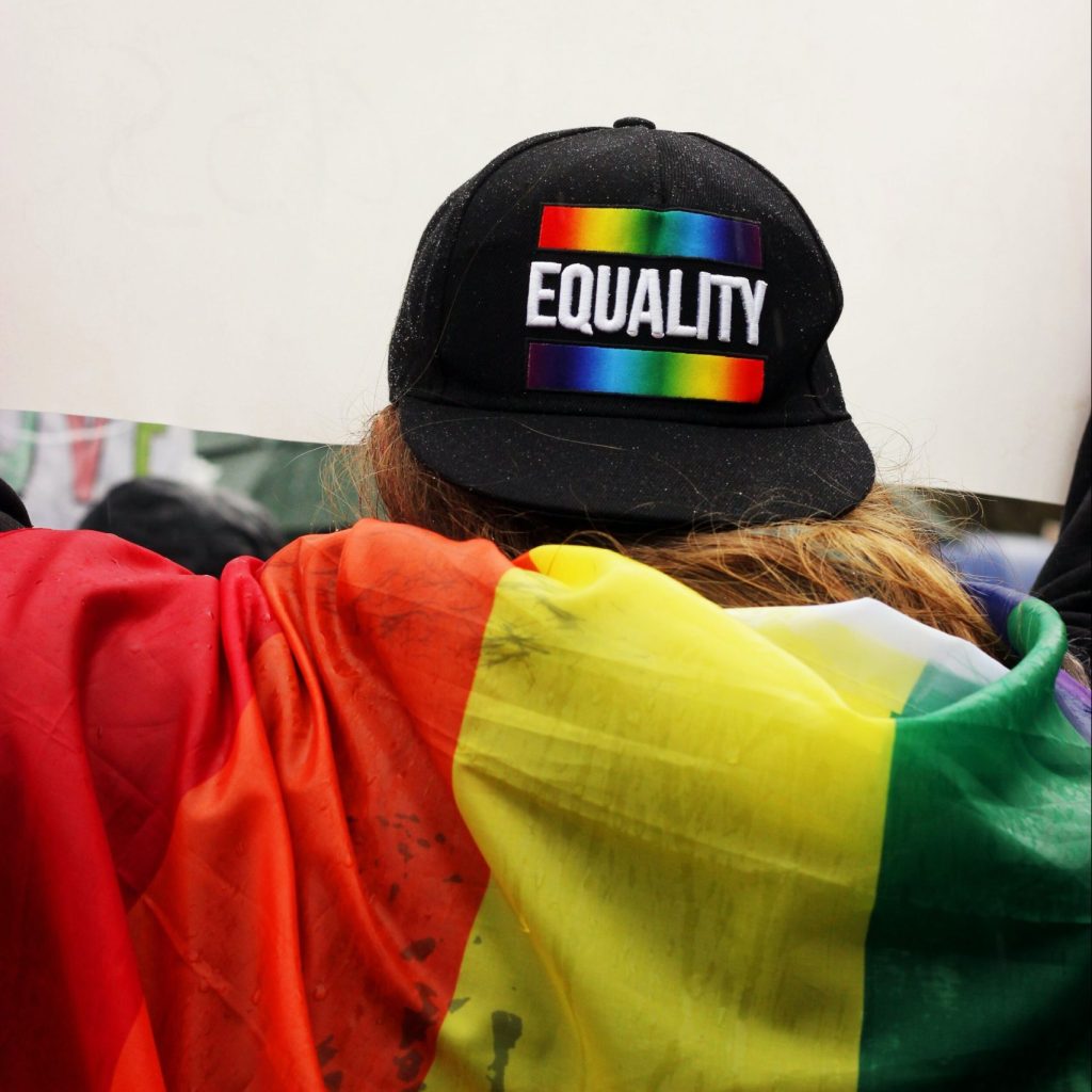 A photograph of someone from behind with a pride flag on their shoulders, wearing a cap backwards with the word "Equality" on
