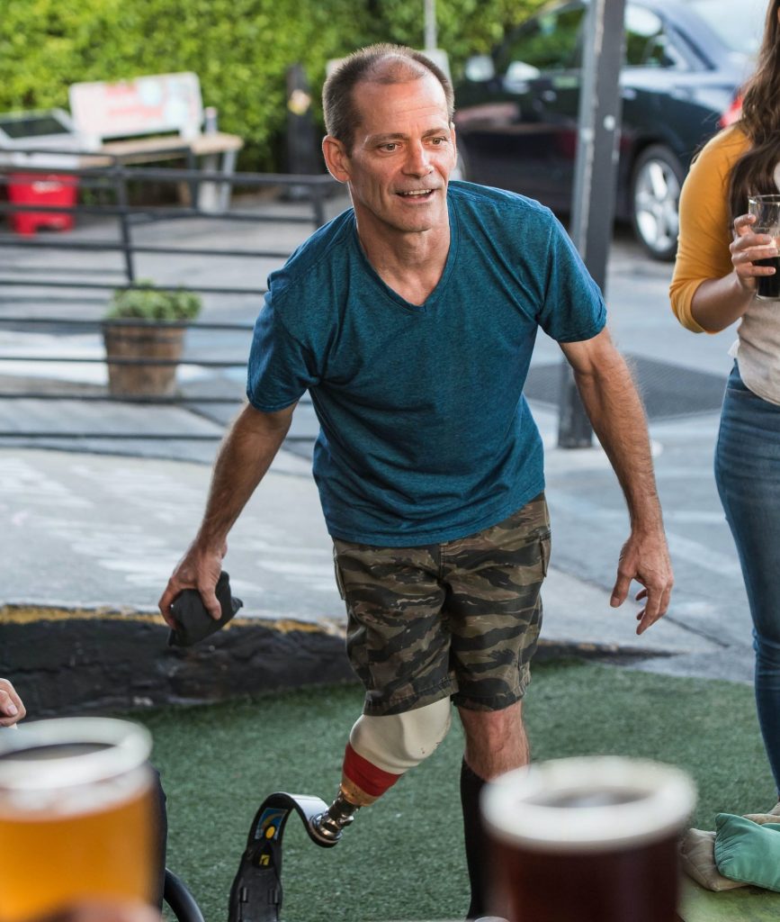 A male with a prosthetic on his left leg, playing a game involving throwing a bean bag,