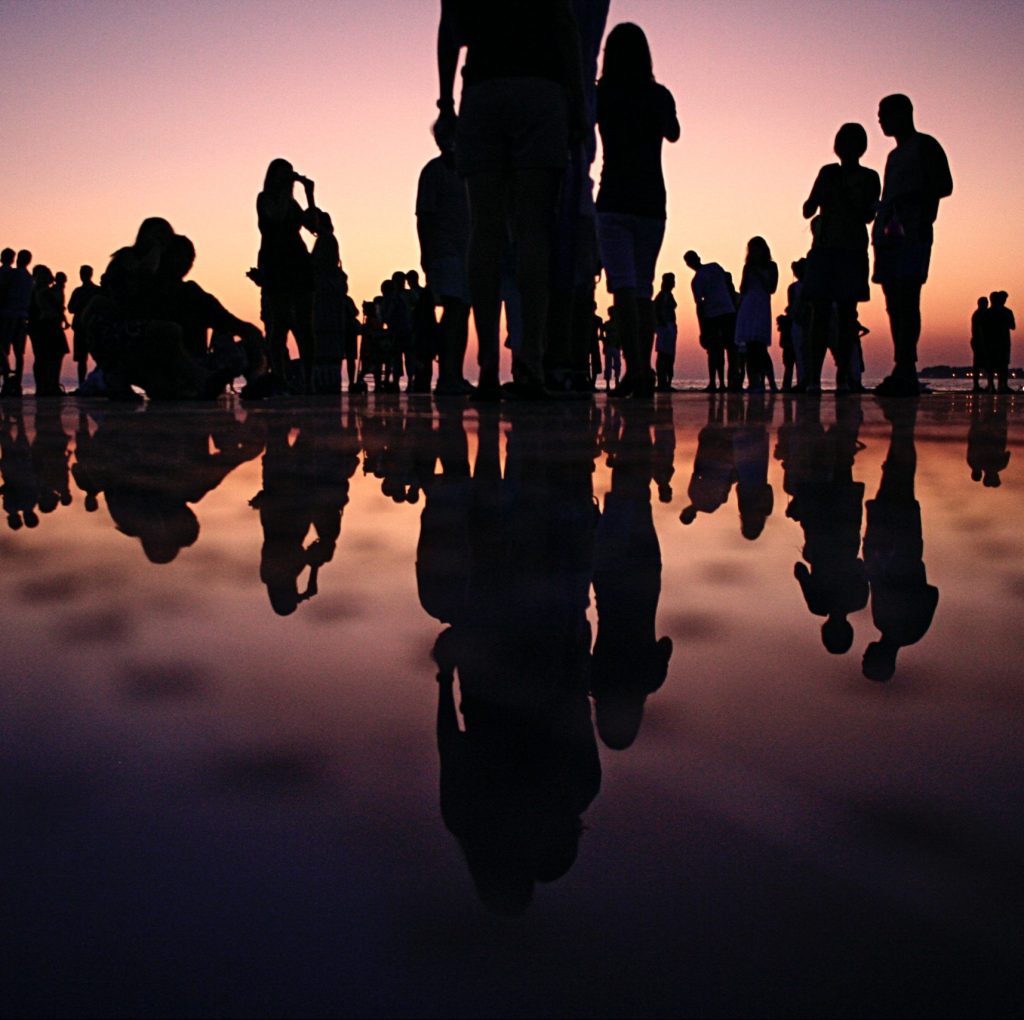 A group of people stood on a beach at sunset, with them silhouetted and their reflections on the sand