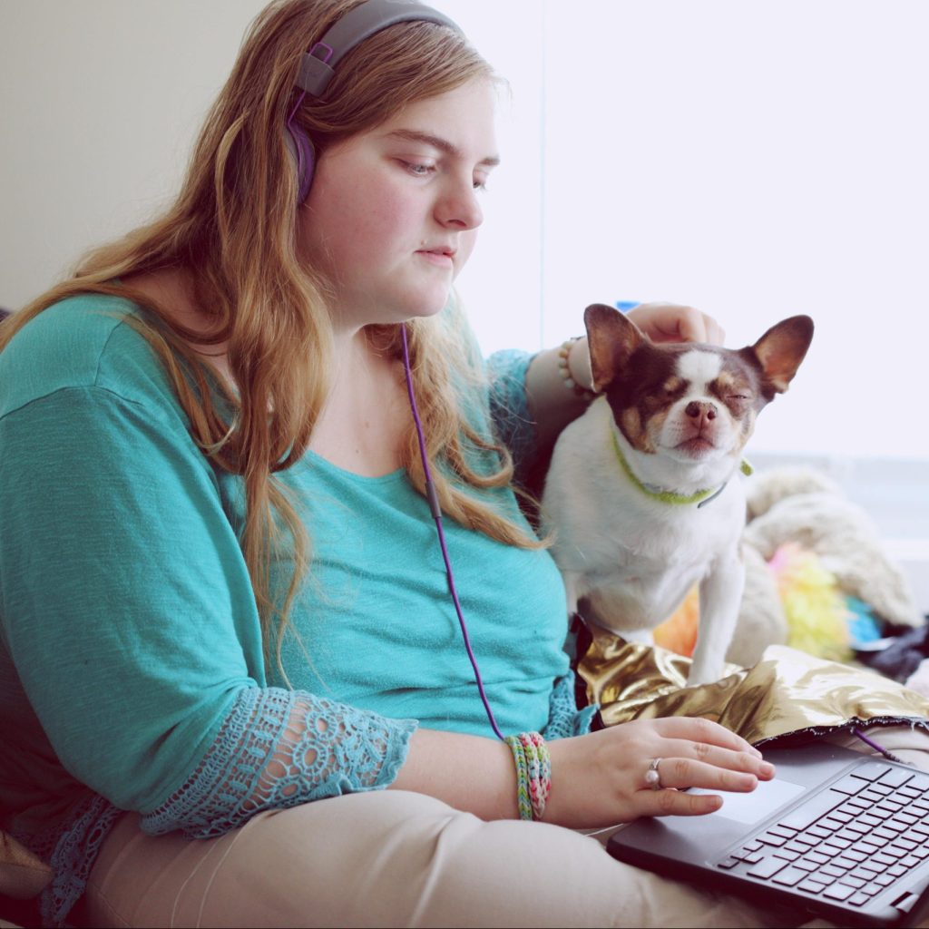 A female sitting on a laptop while wearing over hear headphones and stroking a small dog on the head