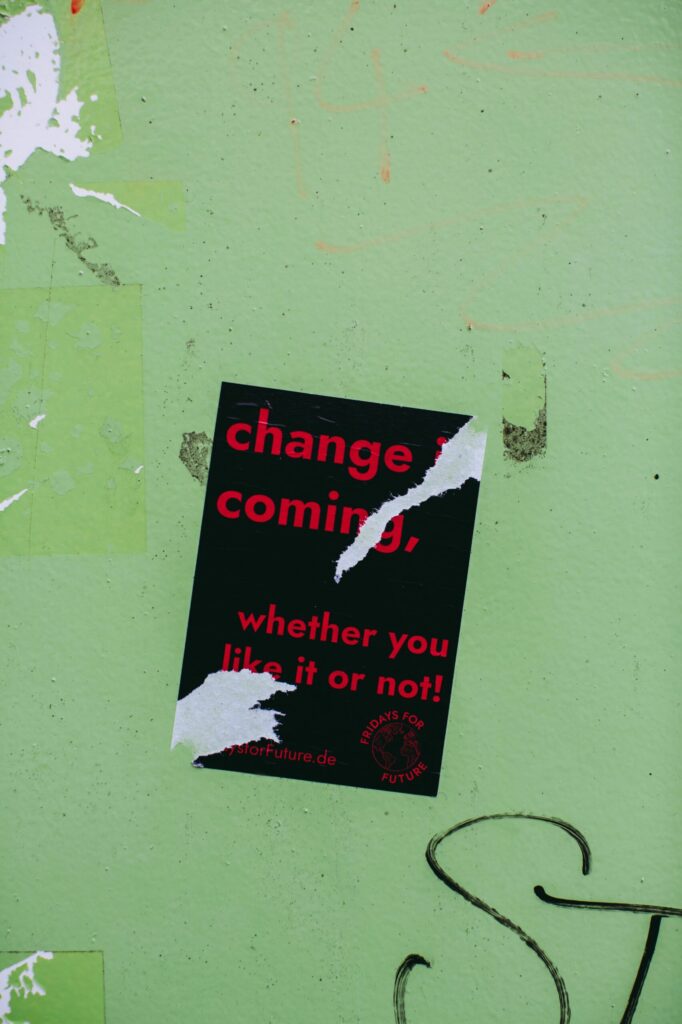 A torn black poster on a green wall with the words 'Change is coming whether you like it or not!' in red.
