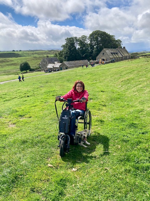 Barbara sits on her bike in open countryside with blue sky and clouds behind her and her small dog by her side